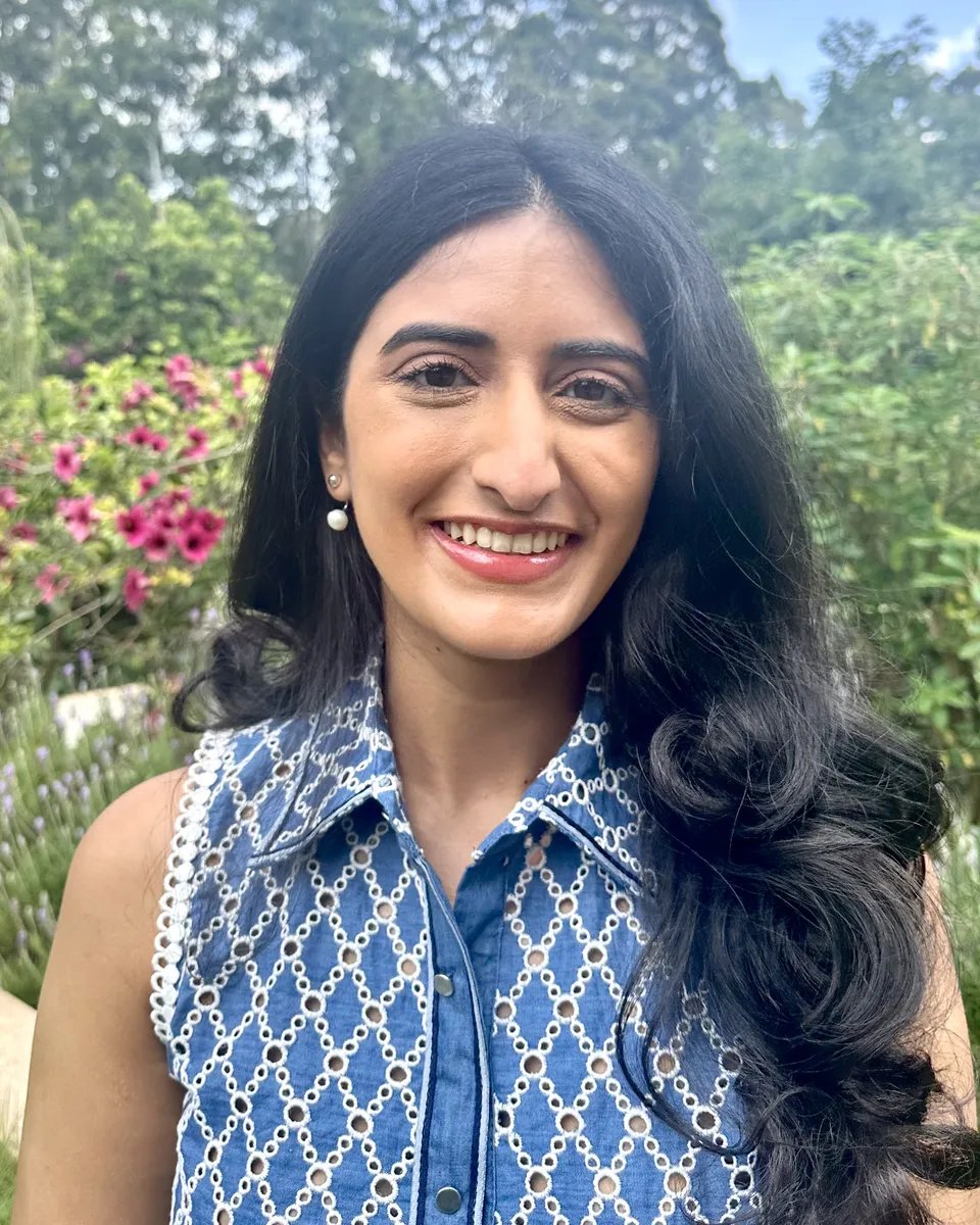 Ashni Dave smiling, with long dark hair, in a blue denim top, standing in a garden.