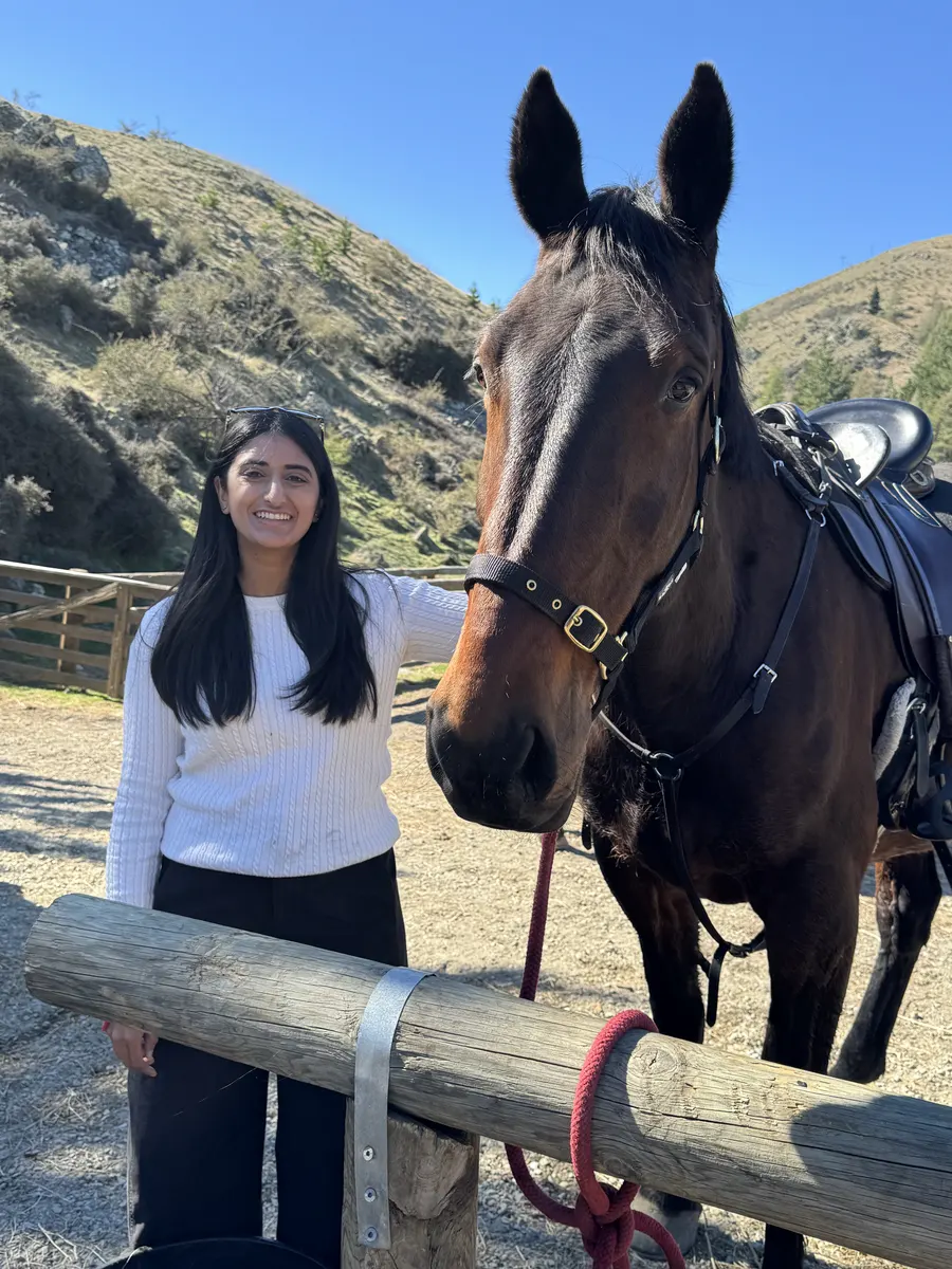 Ashni smiling beside a saddled dark bay horse at a wooden rail, with grassy hills behind.
