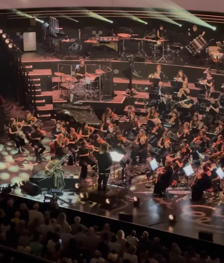 A concert seen from the balcony: an orchestra of strings, percussion and a drum kit on a stepped stage under green beams of light, with the audience in the foreground.