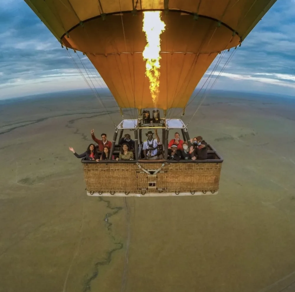 A hot air balloon basket full of waving passengers, seen from above, with its burner flaring over the green plains of the Masai Mara.